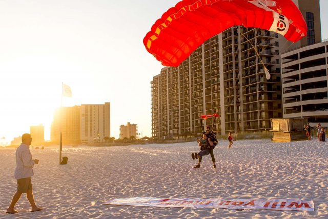 female skydiver landing on the beach right in front of a will you marry me banner laying on the beach